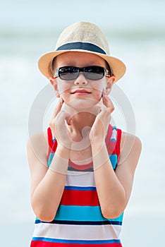 Boy on the beach wearing a hat and glasses smears his skin with