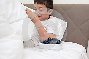 A boy in a bed eating fresh tasty blueberry from a wnite ceramic bowl for breakfast