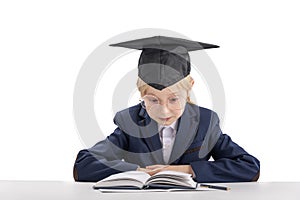 Boy with big glasses wears school uniform and student hat sitting at table reading textbook. Isolated on white background