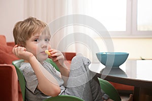 A boy with blon hair is eating corn sticks in the kitchen. Portrait of a cute boy looking at camera in close-up