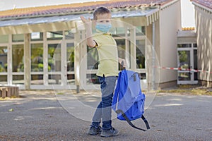 A boy with a blue backpack and wearing a mask waves goodbye in front of the school or kindergarten door. Children are happy to