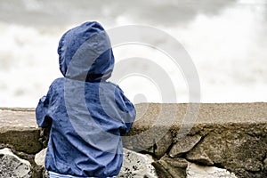 Boy in blue jacket with hood stands with his back against the pier against the background of sea waves