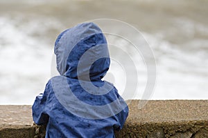 Boy in blue jacket with hood stands with his back against the pier against the background of sea waves
