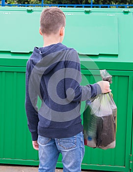 A boy in a blue jumper, carries a full bag of garbage to a container to throw it away, rear view