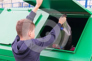A boy in a blue jumper, throws a full bag of garbage into a green container, close-up