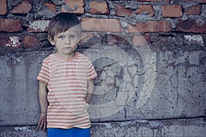 A boy with blue, sad, eyes stands leaning against a brick wall, looking at the camera. Portrait of a beautiful baby.