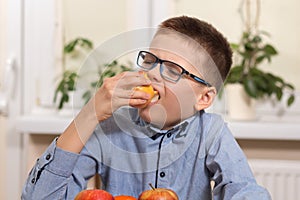 The boy in a blue shirt and glasses sits at the table and holds in his hand an apple which he eats with taste.