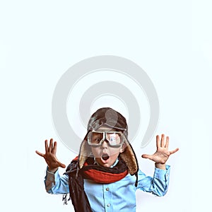 Boy in a blue shirt, pilot`s glasses, hat and scarf raised his hands on a light background