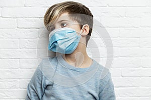 A boy in a blue sweater in a medical mask stands against the background of a white brick wall. Coronavirus pandemic. Close-up