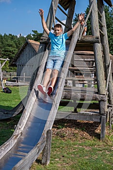 A boy in a blue t-shirt and denim shorts slides down a wooden playground slide, arms raised with exhilaration. Perfect