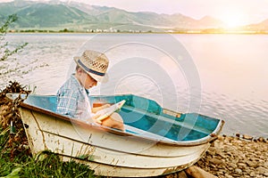 Boy with book seats in old boat on the lake bank
