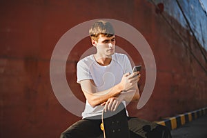 Boy with brown hair sitting with skateboard in hand and thoughtfully looking in cellphone. Young man in white t-shirt