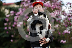 Boy in cap hold cat in hands against nice spring day near magnolia blooming tree