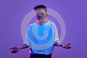 Boy in casual whit t-shirt and glasses spreading hands, standing with misunderstanding face against purple background in