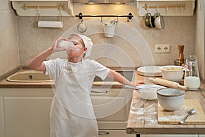 A boy in chef clothes drinks milk from a glass while cooking an apple pie in the kitchen