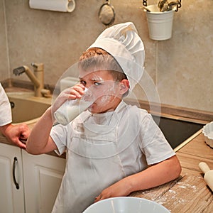 A boy in chef clothes drinks milk from a glass while cooking an apple pie in the kitchen