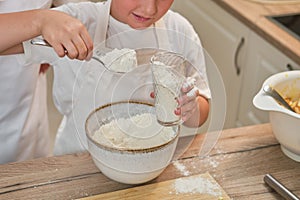 A boy in chef clothes pours flour into the dough for cooking apple pie
