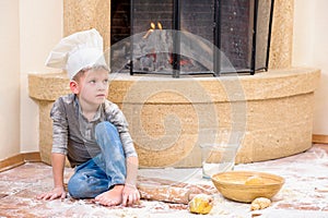 A boy in chef`s hats near the fireplace sitting on the kitchen floor soiled with flour, playing with food, making mess