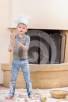 A boy in chef`s hats near the fireplace sitting on the kitchen floor soiled with flour, playing with food, making mess