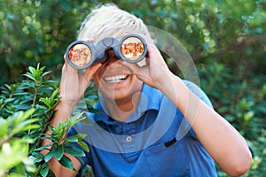 Boy, child with binoculars and search in nature for learning and fun, forest and adventure at summer camp. Young camper