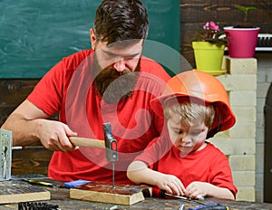 Boy, child busy in protective helmet learning to hammering hobnails with dad. Father, parent with beard teaching little