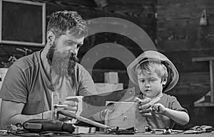 Boy, child busy in protective helmet learning to use handsaw with dad. Father, parent with beard teaching little son to