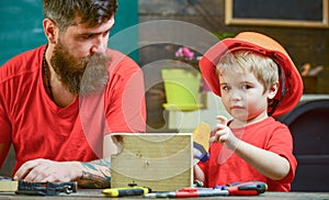 Boy, child busy in protective helmet learning to use handsaw with dad. Father, parent with beard teaching little son to