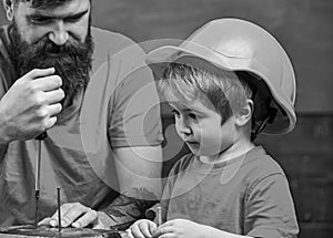 Boy, child busy in protective helmet learning to use screwdriver with dad. Father, parent with beard teaching little son