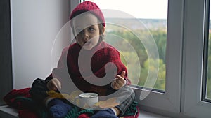 Boy child in a red sweater and a knitted hat sits on a large white window