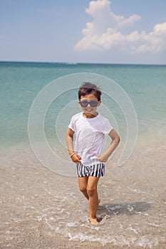 boy child in striped shorts and a white T-shirt walks on sandy beach and in sunglasses