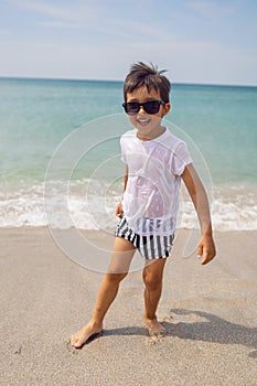 boy child in striped shorts and a white T-shirt walks on sandy beach and in sunglasses