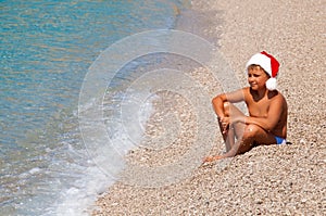Boy in Christmas hat santa claus sitting on the beach