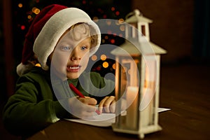 Boy in a Christmas hat writes a letter to Santa Claus by the light of a lantern at the Xmas tree