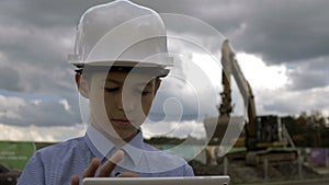 Boy in a construction helmet with a tablet on the background of a combine that digs the ground, the boy is in charge of