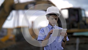 Boy in a construction helmet with a tablet on the background of a combine that digs the ground, the boy is in charge of