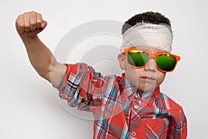 Boy in dark sunglasses, in a plaid shirt, his head is bandaged, checks his muscles, the concept of children`s patience, close-up
