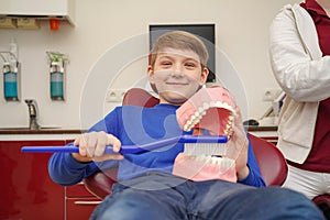 A boy in a doctors chair cleans a plastic jaw with teeth with a large toothbrush