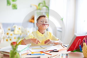 The boy does his homework at home. happy child at the table with school supplies funny smiles and wrinkles nose