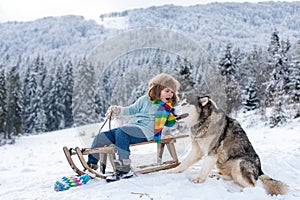 Boy with dog enjoy a sleigh ride. Child sledding, riding a sledge. Children play in snow in winter. Outdoor kids fun for