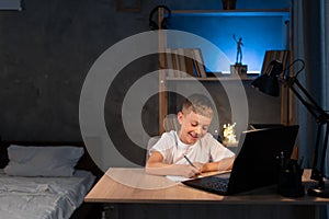 boy is doing homework using a laptop computer at his bedroom desk at night