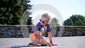 A boy draws drawings with colored crayons on the asphalt, street tiles. A hot summer day.