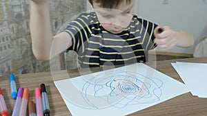 Boy draws spiral of green and orange felt-tip pen on white paper sitting at the table in his room.