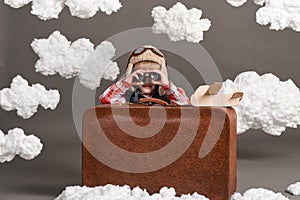 Boy dressed as an airplane pilot sit between the clouds with old suitcase and playing with handmade plane