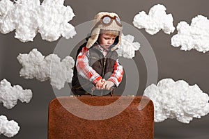 Boy dressed as an airplane pilot stand between the clouds with old suitcase and playing with handmade plane