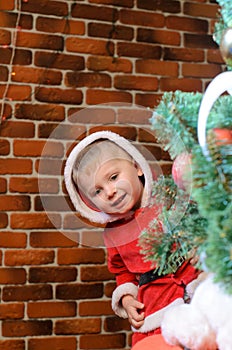 Boy dressed as Santa Claus peeks out and laughs from behind a Christmas tree