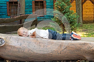 A boy dressed in a white T-shirt and jeans is playing in the park. He lay down on a bench made of a huge tree trunk, he