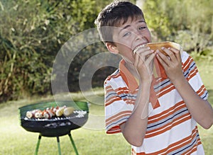 Boy Eating Frankfurter With Barbecue Grill In Background photo