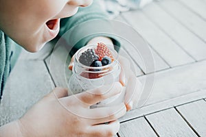 Boy eating Pudding with chia seeds, yogurt and fresh fruits: Strawberries, blueberries and blackberries in glass jars on wooden b