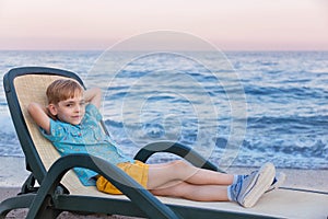 Boy of the European appearance in a blue striped t-shirt of a polo and yellow shorts has a rest in a chaise lounge at a surf strip