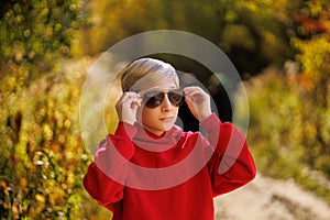 A boy in a fall park corrects his sunglasses. A boy in a red suit in a park in the fall